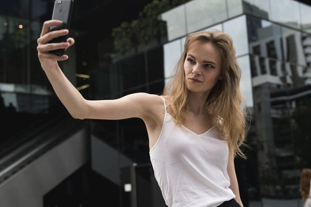 Business woman making selfies in front of office building. Smiling lady taking photos for her business partners abroad. Beautiful brunette business woman photographing selfie on a phone outdoors. - Fotografie, Obrázek