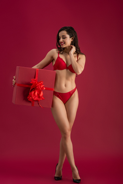 sexy, smiling girl in lingerie and high heeled shoes holding large gift box with ribbon and bow on red background - Photo, image