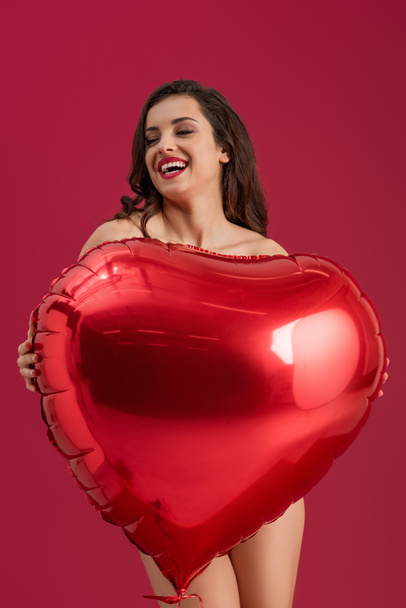 sexy girl smiling with closed eyes while holding large heart-shaped balloon isolated on red - Fotó, kép