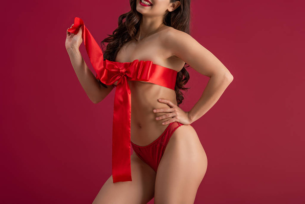 cropped view of sexy girl, wrapped in red satin ribbon with bow, posing isolated on red - Zdjęcie, obraz