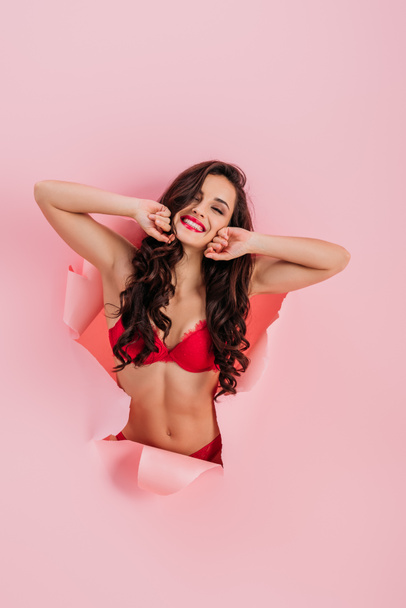 sexy, cheerful girl in red lingerie looking at camera while stretching in paper hole on pink background - Foto, imagen