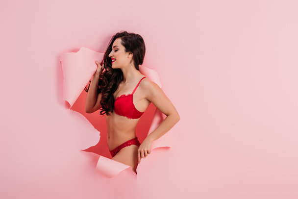 seductive, smiling girl in red lingerie standing in paper hole on pink background - Photo, Image