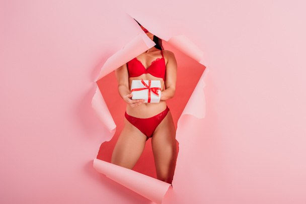 bijgesneden weergave van sexy meisje in lingerie houden geschenk doos terwijl staan in papier gat op roze achtergrond - Foto, afbeelding