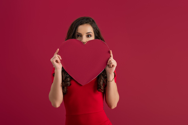 sexy, elegant girl covering face with paper heart while looking at camera isolated on red - Fotografie, Obrázek