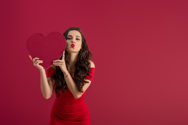 sexy, elegant girl holding paper heart while sending air kiss at camera isolated on red - Photo, image