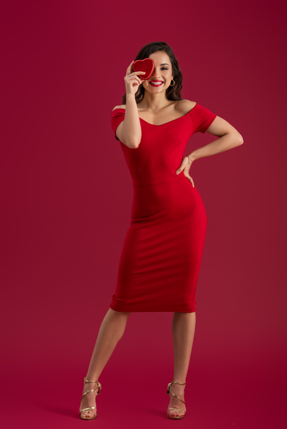 sensual, elegant girl covering eyes with heart-shaped gift box while standing with hand on hip on red background - Fotografie, Obrázek