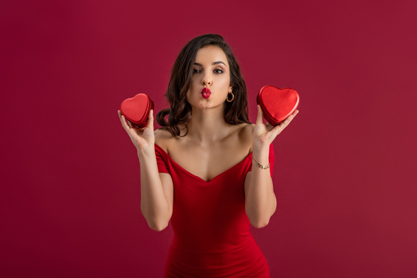σέξι, κομψό κορίτσι που στέλνει φιλί αέρα στην κάμερα κρατώντας ταυτόχρονα κουτιά δώρων σε σχήμα καρδιάς που απομονώνονται στο κόκκινο - Φωτογραφία, εικόνα