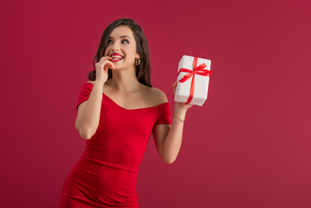dreamy, elegant girl holding gift box while looking away and smiling isolated on red - Photo, Image