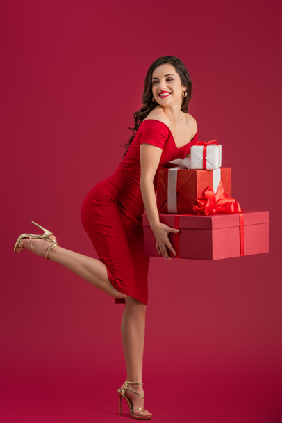 flirty, elegant girl holding gift boxes and looking away while standing on one leg on red background - Photo, image