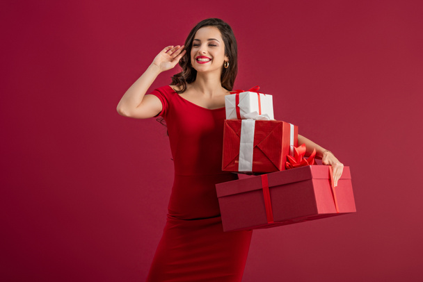 sexy, happy girl looking away and waving hand while holding gift boxes isolated on red - 写真・画像
