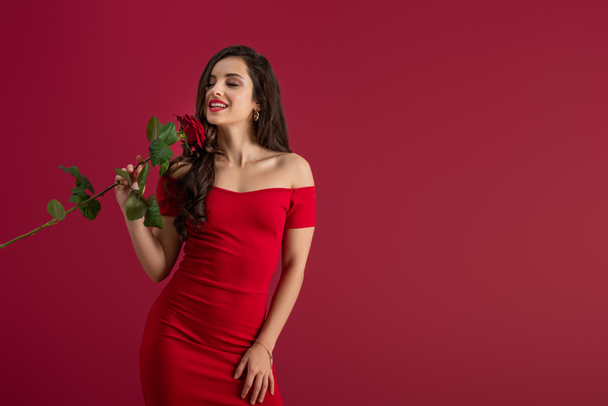 seductive, elegant girl enjoying flavor of red rose with closed eyes isolated on red - Photo, Image