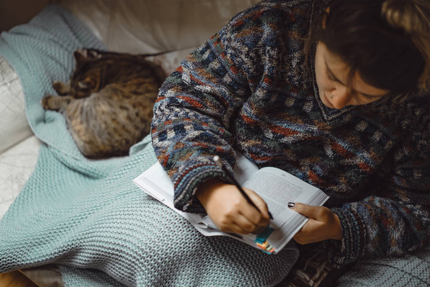 a young woman under a plaid with cat spends a cozy time reading an interesting book. - Foto, Bild