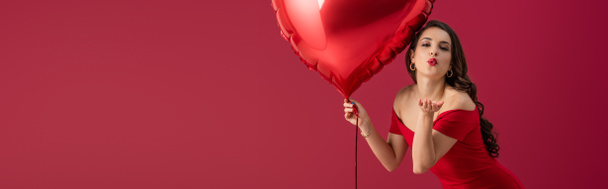 панорамный снимок соблазнительной, элегантной девушки, держащей воздушный шар в форме сердца и посылающей воздушный поцелуй в камеру, изолированную на красном
 - Фото, изображение