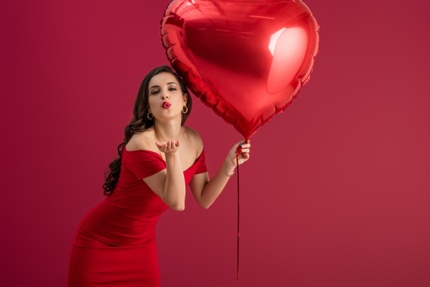 σαγηνευτικό, κομψό κορίτσι κρατώντας μπαλόνι σε σχήμα καρδιάς και στέλνοντας φιλί αέρα στην κάμερα απομονωμένη στο κόκκινο - Φωτογραφία, εικόνα