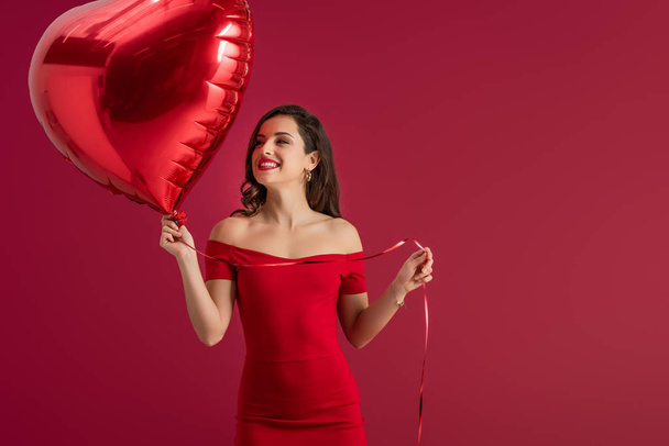 happy, elegant girl looking away while holding heart-shaped balloon isolated on red - Foto, imagen