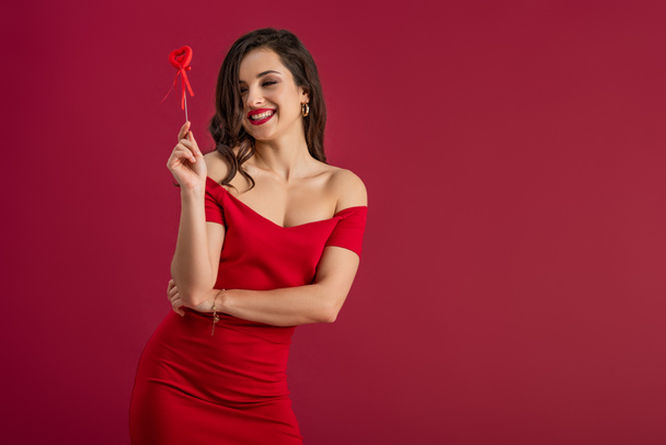 sensuelle, fille élégante tenant coeur jouet sur bâton tout en souriant à la caméra isolée sur rouge
 - Photo, image