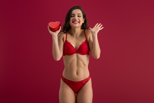 sexy, cheerful girl in lingerie holding heart-shaped gift box while standing with closed eyes isolated on red - Foto, afbeelding