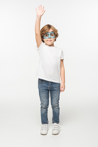 full length view of smiling boy with superhero mask painted on face standing with raised hand on white background - Photo, Image