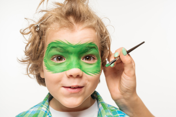 visão recortada do artista pintando máscara de gecko no rosto de menino desgrenhado e alegre isolado no branco
 - Foto, Imagem