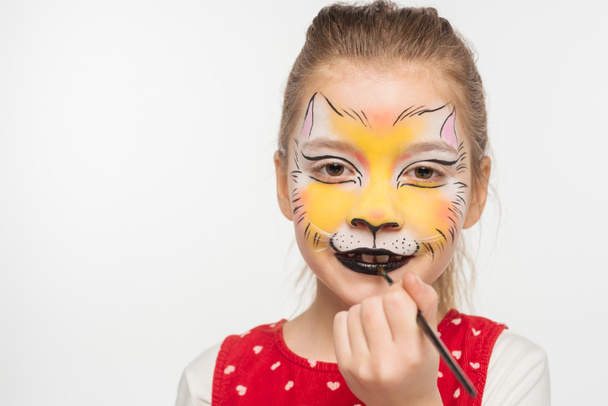 cute kid with painted tiger muzzle on face painting on lips with paintbrush isolated on white - Fotografie, Obrázek