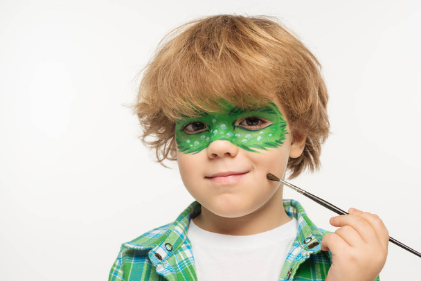 menino bonito com máscara de gecko pintado no rosto tocando bochecha com pincel isolado no branco
 - Foto, Imagem