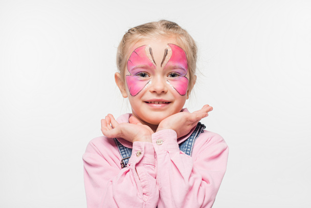 lächelndes Kind mit Schmetterlingsmalerei im Gesicht, die Hände vor dem Gesicht haltend, während es isoliert auf weiß in die Kamera schaut - Foto, Bild