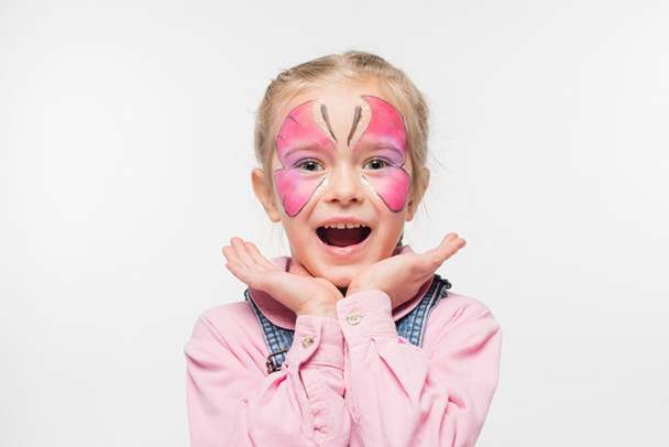 excited child with butterfly painting on face holding hands near face while looking at camera isolated on white - Fotó, kép
