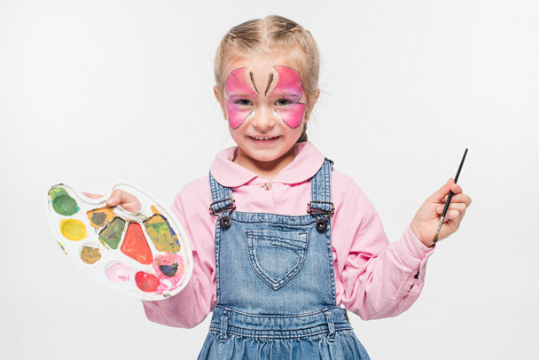 smiling child with butterfly painting on face holding palette and paintbrush while looking at camera isolated on white - 写真・画像