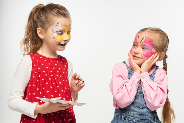 cute child with cat muzzle painting on face holding palette and paintbrush near amused friend with painted butterfly mask isolated on white - Photo, Image