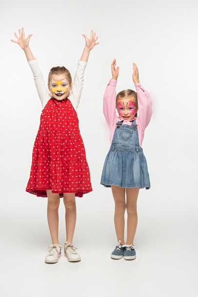 full length view of adorable children with cat muzzle and butterfly paintings on faces standing with raised hands on white background - Zdjęcie, obraz
