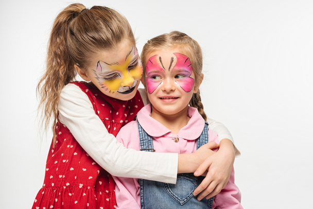 adorable kid with cat muzzle painting on face embracing friend with painted butterfly mask isolated on white - 写真・画像