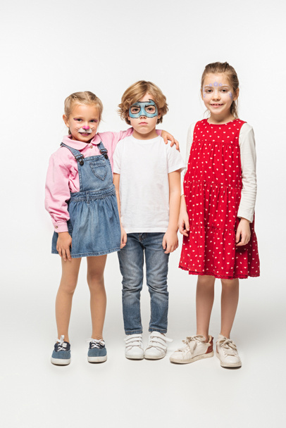 full length view of cheerful kids and offended boy with colorful face paintings looking at camera on white background - Photo, Image