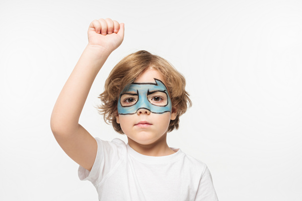 serious boy with superhero mask painted on face standing with raised fist while looking at camera isolated on white - Fotoğraf, Görsel