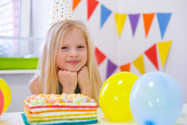 Blonde caucasian girl sits thoughtfully and dreamily at festive table near birthday rainbow cake and makes a wish. looking at camera. Colorful background with balloons - Fotografie, Obrázek