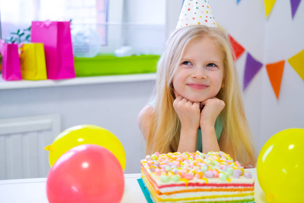 Blonde caucasian girl sits thoughtfully and dreamily at festive table near birthday rainbow cake and makes a wish. Colorful background with balloons - Foto, imagen