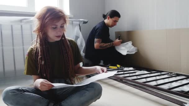 Young couple of hipsters in new home to assemble furniture. Tattooed male and redhead female with dreadlocks unpacking new bed. - Materiał filmowy, wideo