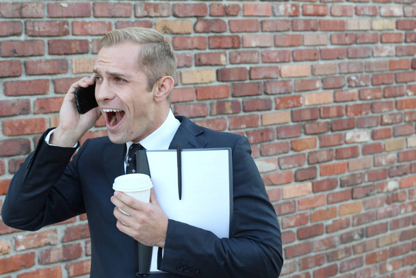 primer plano de hombre de negocios adulto guapo con carpeta y taza de papel de café hablando por teléfono en frente de la pared de ladrillo en la calle
 - Foto, imagen