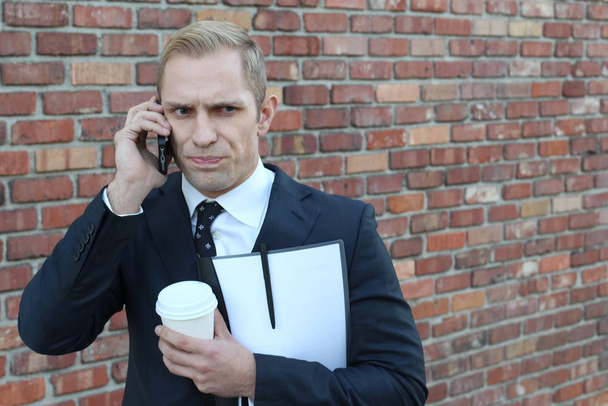 közeli kép jóképű felnőtt üzletember mappával és papír csésze kávé beszél telefonon előtt téglafal az utcán - Fotó, kép