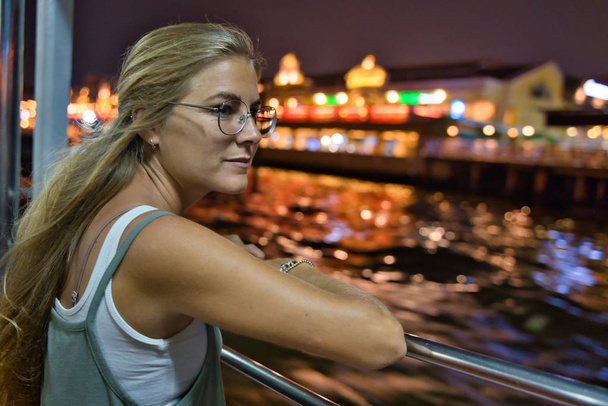 Blondynka z okularami patrząca na rzekę opierająca się na żelaznej poręczy w nocy ze światłami budynków w Tajlandii - Zdjęcie, obraz