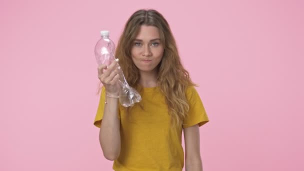 Irritated caucasian woman in yellow t-shirt is holding plastic bottles while crumpling them isolated over pink background - Кадры, видео