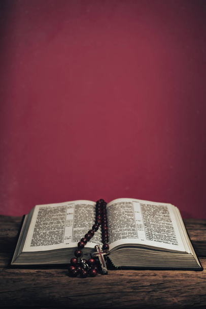 Avaa Pyhä Raamattu ja helmiä krusifiksi vanhan tammen puupöydällä. Kaunis punainen seinä tausta
. - Valokuva, kuva
