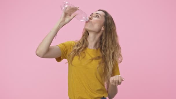 A pretty young woman drinks water from plastic bottle then throws it to the side isolated over pink background - Séquence, vidéo