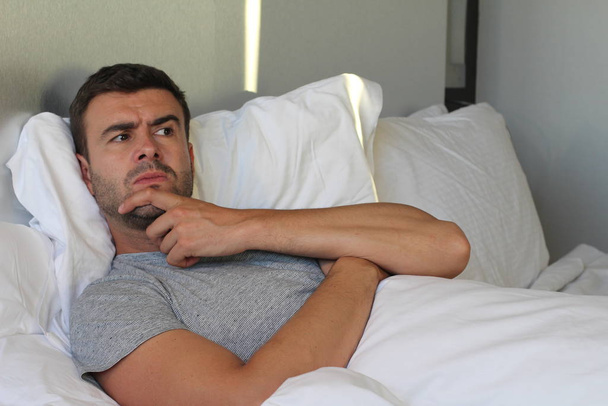 zblízka portrét pohledného mladého expresivního muže dotýkajícího se brady doma v posteli - Fotografie, Obrázek