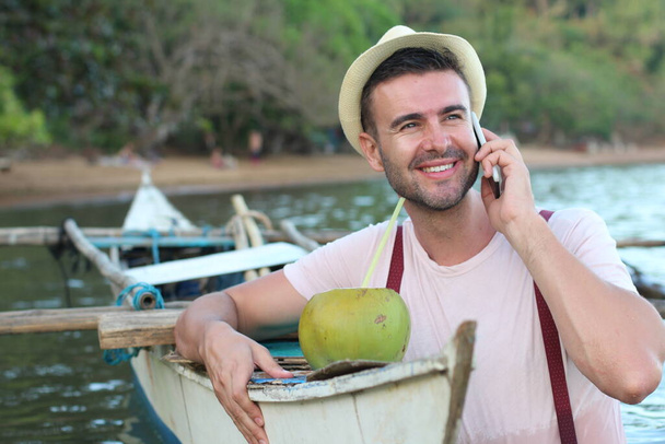 Stylish male calling by phone from a boat while drinking a fresh coconut - Photo, Image