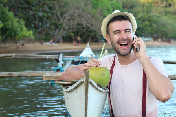 Κομψό αρσενικό καλώντας από το τηλέφωνο από ένα σκάφος, ενώ πίνοντας μια φρέσκια καρύδα - Φωτογραφία, εικόνα