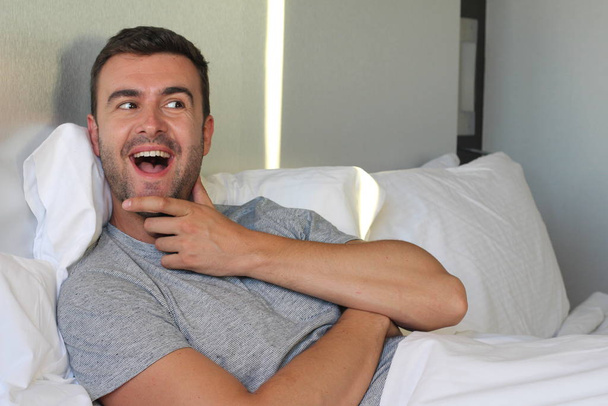 portrait de près d'un beau jeune homme expressif qui se touche le menton dans son lit à la maison - Photo, image