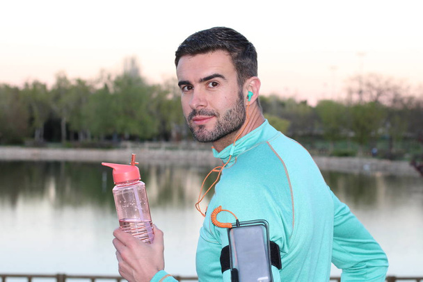 крупним планом портрет красивого молодого бігуна зі смартфоном, спортивною пляшкою та навушниками перед озером в парку
 - Фото, зображення