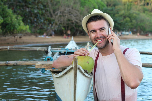 Stylish male calling by phone from a boat while drinking a fresh coconut - Photo, Image