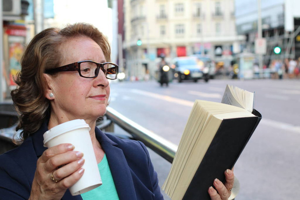 Ώριμη γυναίκα πίνοντας καφέ και διαβάζοντας βιβλίο κάθεται σε εσωτερικούς χώρους σε αστικό καφέ. Cafe τρόπο ζωής της πόλης με φανάρια στο παρασκήνιο - Φωτογραφία, εικόνα