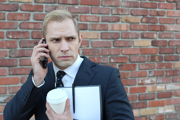 close-up shot of handsome adult businessman with folder and paper cup of coffee talking by phone in front of brick wall on street - Photo, Image